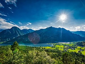 Eine bergige Landschaft mit grünen Wiesen, einem See und einem bewölkten, sonnigen Himmel.